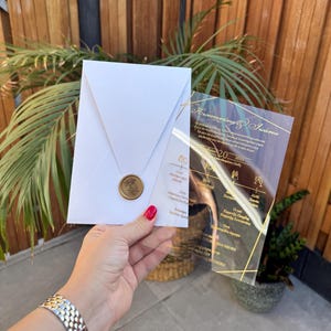Puede incluir: Un sobre blanco con un sello dorado y una invitación de acrílico transparente con texto y diseño dorado. La invitación muestra la fecha, hora y lugar de un evento.