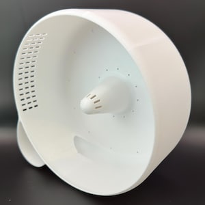 Könnte beinhalten: Ein weißes, kreisförmiges Kunststoffgerät mit einem kegelförmigen Mittelteil und einer Reihe kleiner Löcher. Das Gerät hat ein gebogenes, offenes Design mit rechteckigen Lüftungsschlitzen an der Seite. Das Objekt befindet sich auf einer dunklen Oberfläche.