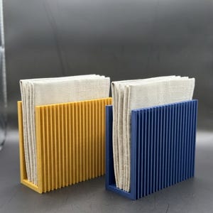 K&ouml;nnte beinhalten: Zwei Serviettenhalter, einer in Senfgelb und einer in Marineblau, die jeweils einen Stapel beiger Leinen-Servietten halten. Die Halter haben ein vertikales Rippendesign und eine rechteckige Form, ideal f&uuml;r einen Esstisch.