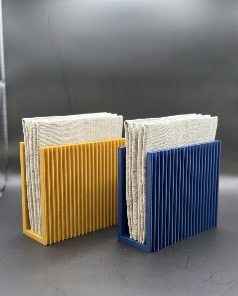 K&ouml;nnte beinhalten: Zwei Serviettenhalter, einer in Senfgelb und einer in Marineblau, die jeweils einen Stapel beiger Leinen-Servietten halten. Die Halter haben ein vertikales Rippendesign und eine rechteckige Form, ideal f&uuml;r einen Esstisch.