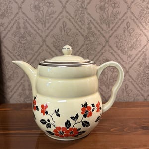 May include: Cream-coloured ceramic teapot with a lid and handle. The teapot features a black and red floral design around the base and a silver trim. The teapot is sitting on a wooden surface.