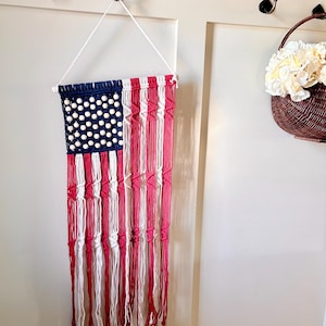 May include: A macrame wall hanging designed to resemble the American flag, featuring blue, white, and red cotton cord. The blue section includes light-colored wooden beads, while the red and white sections are created with knotted cord patterns.