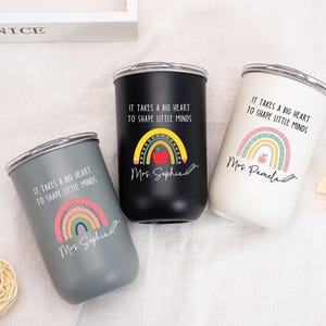 May include: Three stainless steel tumblers in gray, black, and white. Each features a rainbow graphic, a pencil, an apple, and the text "IT TAKES A BIG HEART TO SHAPE LITTLE MINDS" along with personalized names "Mrs. Sophia" or "Mrs. Pamela" in script.