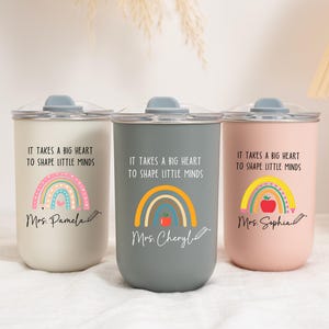 May include: Three cylindrical tumblers in cream, gray, and light pink, each with a clear lid. The text "IT TAKES A BIG HEART TO SHAPE LITTLE MINDS" is printed above a rainbow graphic, with personalized names and a pencil icon at the bottom.