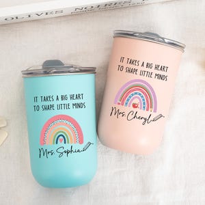 May include: Two stainless steel tumblers with lids, one light blue and one light pink. Each features a colorful rainbow graphic, a small apple icon, a pencil illustration, and the text "IT TAKES A BIG HEART TO SHAPE LITTLE MINDS" with names.