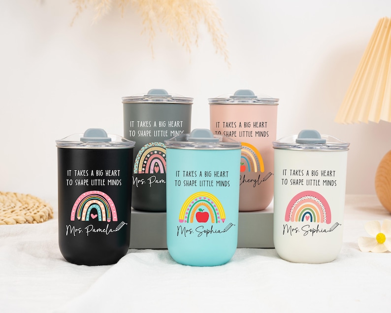 May include: Five cylindrical tumblers with lids in black, dark gray, light blue, pink, and white. Each features a rainbow graphic, a pencil, and the text "It takes a big heart to shape little minds" along with a personalized name in script font.