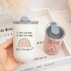 May include: A white tumbler and a smaller pink tumbler, both featuring a clear lid with a grey stopper. Each cup displays a colorful rainbow graphic, a pencil icon, and the text "IT TAKES A BIG HEART TO SHAPE LITTLE MINDS" with a name printed below.