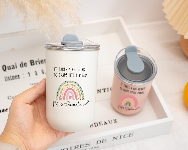 May include: A white tumbler and a smaller pink tumbler, both featuring a clear lid with a grey stopper. Each cup displays a colorful rainbow graphic, a pencil icon, and the text "IT TAKES A BIG HEART TO SHAPE LITTLE MINDS" with a name printed below.