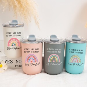 May include: Four cylindrical tumblers with clear lids and grey stoppers in white, pink, dark grey, and teal. Each features a rainbow graphic, the text "IT TAKES A BIG HEART TO SHAPE LITTLE MINDS," and a personalized name written in cursive script.