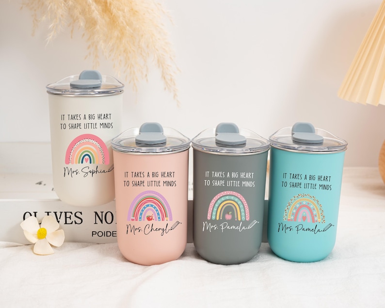 May include: Four cylindrical tumblers with clear lids and grey stoppers in white, pink, dark grey, and teal. Each features a rainbow graphic, the text "IT TAKES A BIG HEART TO SHAPE LITTLE MINDS," and a personalized name written in cursive script.