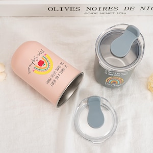 May include: A light pink tumbler and a grey tumbler, each with a clear lid featuring a grey sliding closure. The pink tumbler displays a rainbow, an apple, and the text "It takes a big heart to shape little minds" with "Mrs. Sophia".