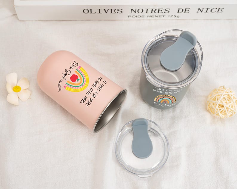 May include: A light pink tumbler and a grey tumbler, each with a clear lid featuring a grey sliding closure. The pink tumbler displays a rainbow, an apple, and the text "It takes a big heart to shape little minds" with "Mrs. Sophia".