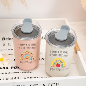 May include: Two stainless steel tumblers with clear lids and grey stoppers. One is light pink, the other white. Both feature the text "IT TAKES A BIG HEART TO SHAPE LITTLE MINDS" and a rainbow graphic. The pink one says "Mrs. Cheryl" and the white one "Mrs. Sophia".