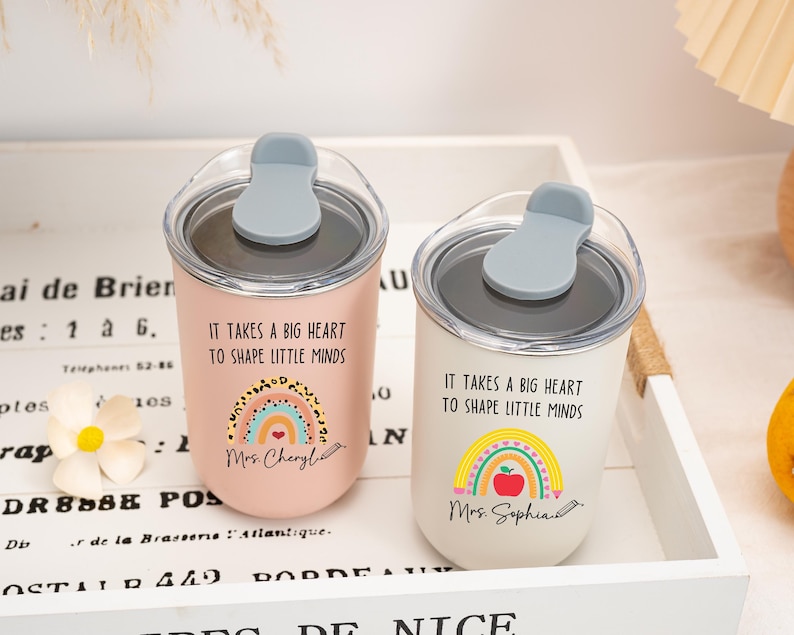 May include: Two stainless steel tumblers with clear lids and grey stoppers. One is light pink, the other white. Both feature the text "IT TAKES A BIG HEART TO SHAPE LITTLE MINDS" and a rainbow graphic. The pink one says "Mrs. Cheryl" and the white one "Mrs. Sophia".