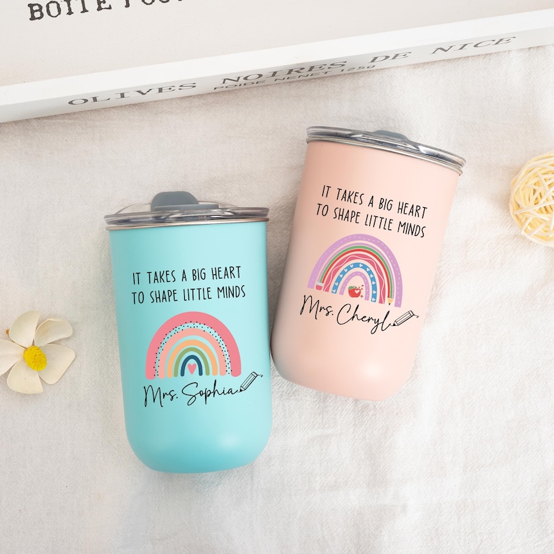 May include: Two stainless steel tumblers with lids, one light blue and one light pink. Each features a colorful rainbow graphic, a small apple icon, a pencil illustration, and the text "IT TAKES A BIG HEART TO SHAPE LITTLE MINDS" with names.