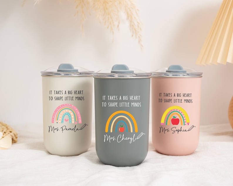 May include: Three cylindrical tumblers in cream, gray, and light pink, each with a clear lid. The text "IT TAKES A BIG HEART TO SHAPE LITTLE MINDS" is printed above a rainbow graphic, with personalized names and a pencil icon at the bottom.