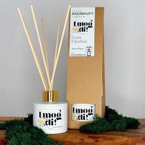 Könnte beinhalten: Eine weiße Diffuserflasche mit goldenem Deckel und Stäbchen, beschriftet mit "Imod tli!" mit einem Duft von Zitrone und Basilikum. Die Flasche steht auf einer Holzoberfläche mit grünem Moos. Das Produkt ist in einem braunen Karton mit dem gleichen Etikett verpackt.