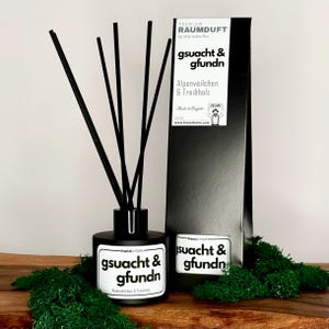 Könnte beinhalten: Schwarze Diffuserflasche mit schwarzen Ruten und einem weißen Etikett mit der Aufschrift "gsuacht & gfundn Alpenveilchen & Treibholz". Die Flasche steht auf einem Bett aus grünem Moos.