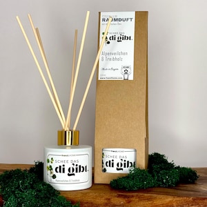 Könnte beinhalten: Weiße Diffuserflasche mit goldenem Deckel und Holzstäbchen. Die Flasche hat ein Etikett mit der Aufschrift "SCHEE DAS di gibt." und "Alpenveilchen & Treibholz". Die Flasche steht auf einem Bett aus grünem Moos.