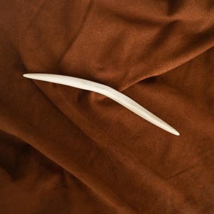 Pode incluir: Um palito de cabelo na cor branco-osso, liso e com um design curvo e cônico. O objeto apresenta uma superfície sólida e polida que se estreita em ponta em ambas as extremidades, exibindo uma forma simples e alongada sem adornos.