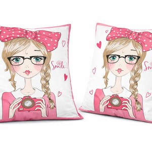 May include: Two decorative throw pillows with a cartoon illustration of a girl with blonde braids, glasses, and a pink bow. She holds a camera. The pillows have a white background with pink hearts and trim. The word "Smile" is written on the pillow.