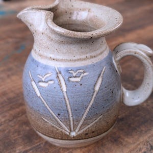 May include: A ceramic pitcher with a light blue and brown color scheme. The pitcher features a handle and a spout. The design includes stylized cattails and birds. The pitcher is likely handmade and has a rustic aesthetic.