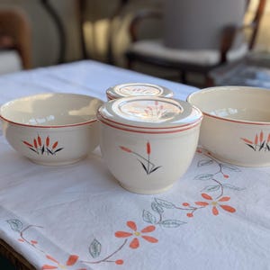 May include: A set of vintage cream-colored ceramic bowls and a lidded container. Each piece features a red rim and a stylized floral design in black, red, and orange. The bowls are arranged on a white tablecloth with a floral pattern.