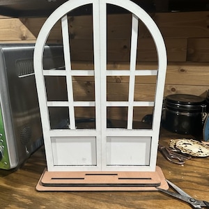 May include: A white wooden window frame with an arched top and a rectangular base. The window has a grid pattern and two lower panels. The frame is set on a light brown base.