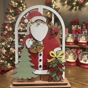 May include: Wooden Christmas decoration featuring Santa Claus in a red suit, white beard, and hat. The design includes a green Christmas tree, bells, and a bow. The decoration is set on a wooden base, with a Christmas tree and snow globes in the background.