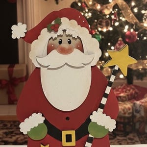 May include: Wooden Santa Claus figure with a red suit, white beard, and a red hat with holly. He holds a star-topped wand. The figure has a black belt with a gold buckle and three gold stars on the front. A festive Christmas decoration.