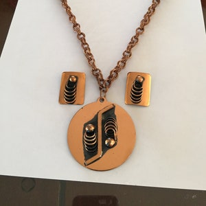 May include: A copper-toned necklace and earring set. The necklace features a chain and a pendant with a circular base and geometric design. The earrings are rectangular with matching details. The set has a vintage aesthetic.