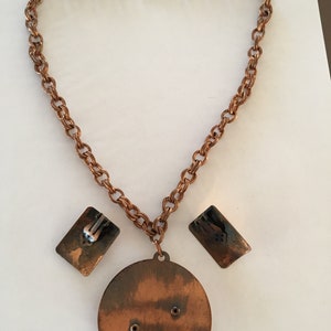 May include: A vintage copper-toned jewelry set featuring a necklace and matching earrings. The necklace has a chain with a large circular pendant and rectangular earrings. The set is displayed on a white surface.