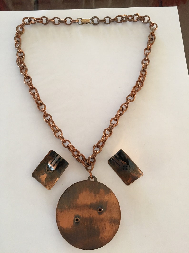 May include: A vintage copper-toned jewelry set featuring a necklace and matching earrings. The necklace has a chain with a large circular pendant and rectangular earrings. The set is displayed on a white surface.