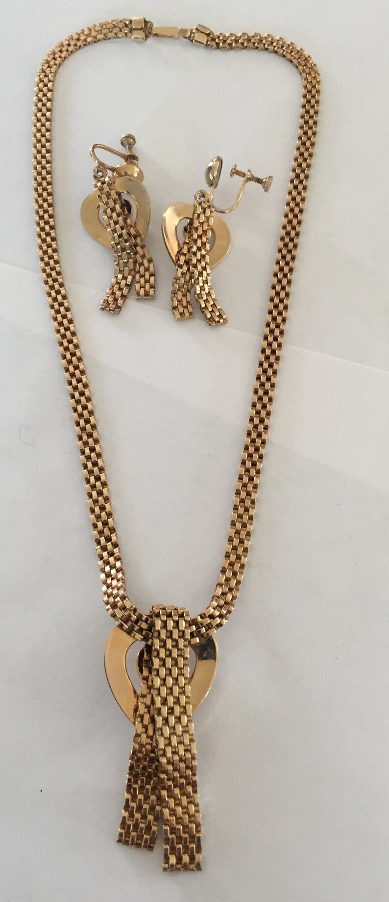 May include: A gold tone necklace and earrings set with a heart-shaped pendant and matching heart-shaped earrings. The necklace and earrings are made of a chain link design.