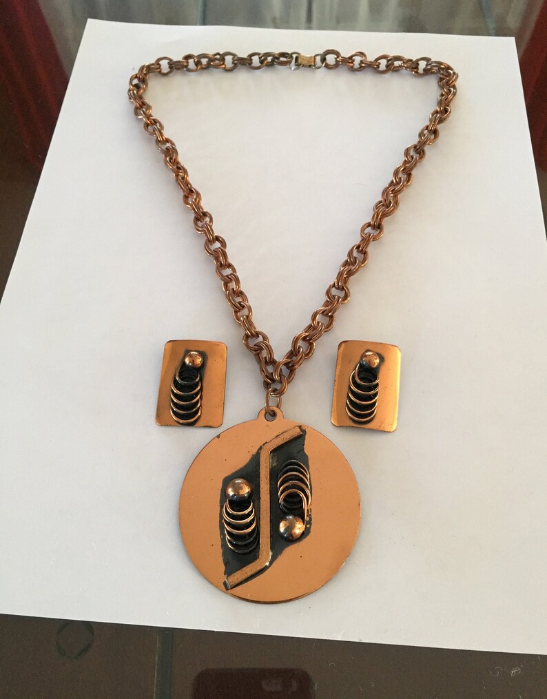 May include: A copper-toned necklace and earring set. The necklace features a chain and a pendant with a circular base and geometric design. The earrings are rectangular with matching details. The set has a vintage aesthetic.