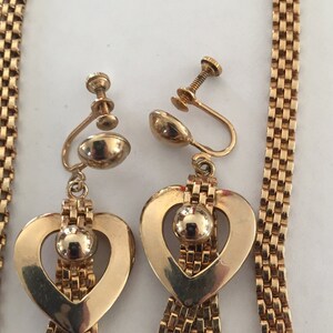 May include: A gold tone necklace and matching earrings with a heart design and a dangling chain. The heart has a round bead in the center.