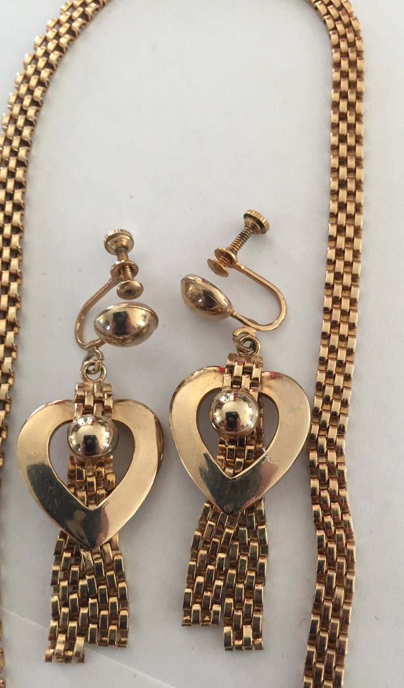 May include: A gold tone necklace and matching earrings with a heart design and a dangling chain. The heart has a round bead in the center.