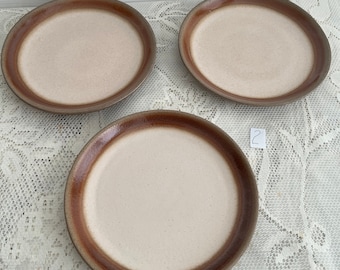 May include: Three brown and white ceramic plates with a brown rim. The plates are on a white lace tablecloth.