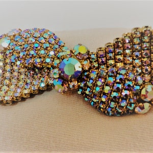 May include: A gold-toned brooch with iridescent rhinestones arranged in a swirling pattern. The brooch is made of many small rhinestones that create a shimmering effect.