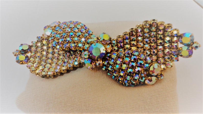 May include: A gold-toned brooch with iridescent rhinestones arranged in a swirling pattern. The brooch is made of many small rhinestones that create a shimmering effect.