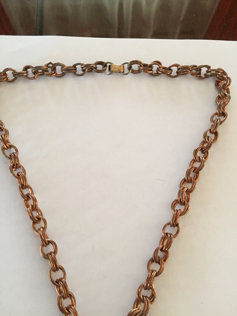 May include: A copper-toned necklace with a double-link chain design. The necklace features a gold-colored clasp. The chain is made of small, interconnected oval links, creating a textured appearance. The necklace is displayed against a white background.