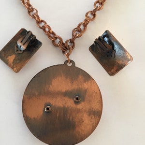 May include: Vintage copper-toned jewelry set featuring a necklace with a round pendant and matching clip-on earrings. The necklace has a chain link design. The earrings are rectangular. The set has a rustic, aged appearance.