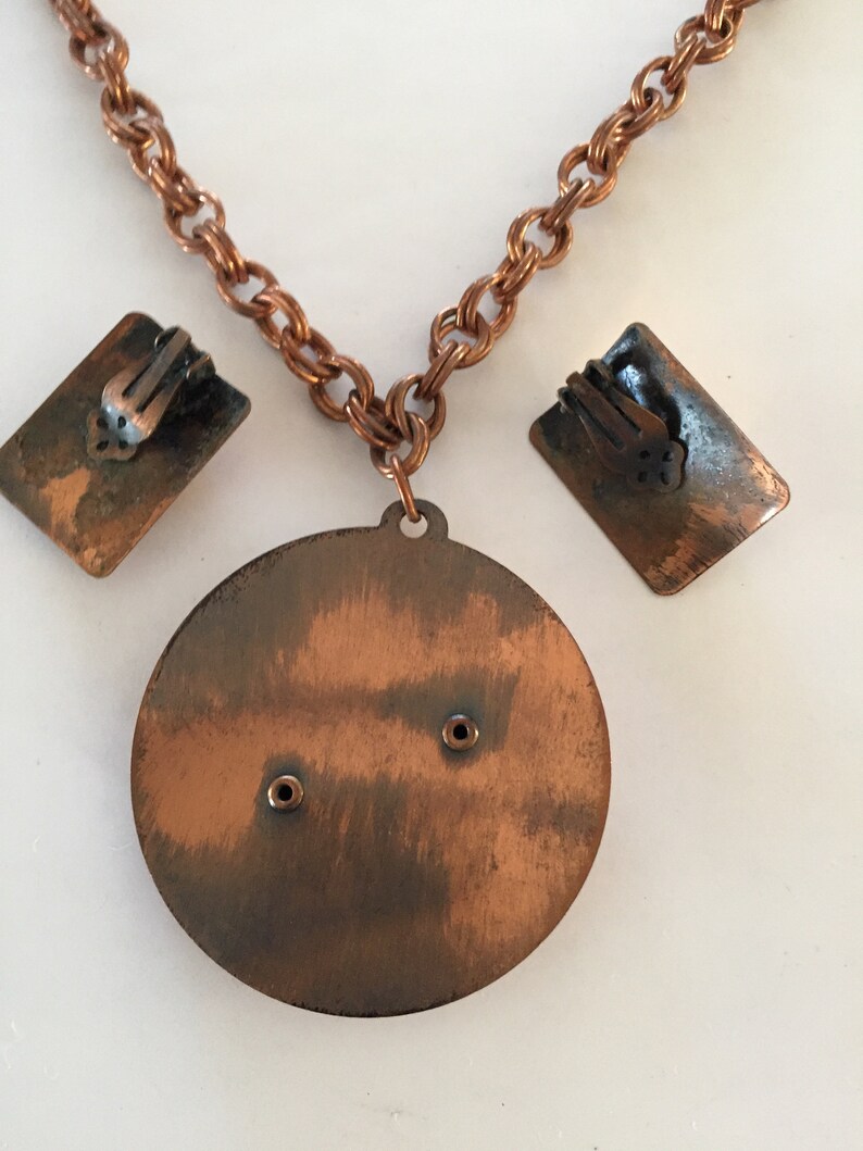 May include: Vintage copper-toned jewelry set featuring a necklace with a round pendant and matching clip-on earrings. The necklace has a chain link design. The earrings are rectangular. The set has a rustic, aged appearance.