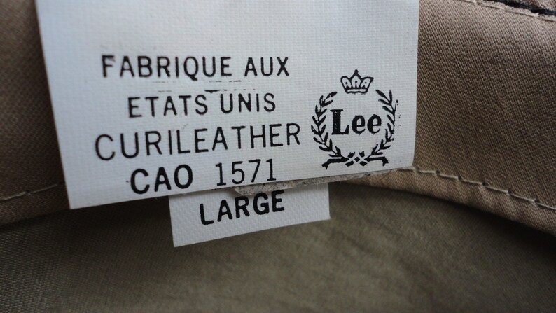 May include: A white tag with black text that reads "Fabrique aux Etats Unis Curileather CAO 1571 Large" and a Lee logo with a crown and wreath.