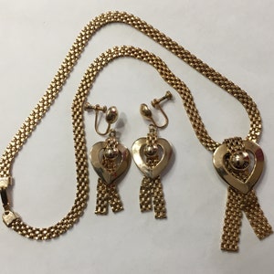 May include: A gold necklace and earrings set with a heart-shaped pendant and dangling chains. The necklace is made of a chain link design and the earrings are clip-on.
