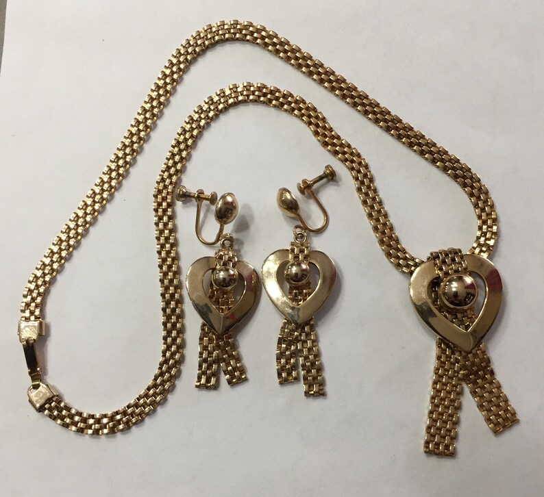May include: A gold necklace and earrings set with a heart-shaped pendant and dangling chains. The necklace is made of a chain link design and the earrings are clip-on.
