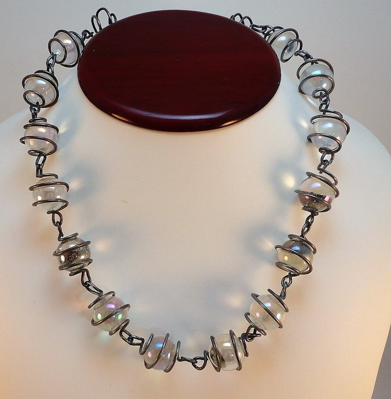 May include: A silver necklace with iridescent glass beads wrapped in wire. The beads are round and have a subtle rainbow effect.