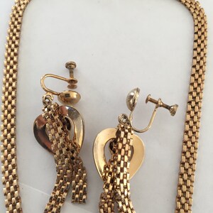 May include: A gold tone necklace and matching earrings with a heart-shaped pendant and dangling chains. The necklace is a box chain and the earrings are clip-on.
