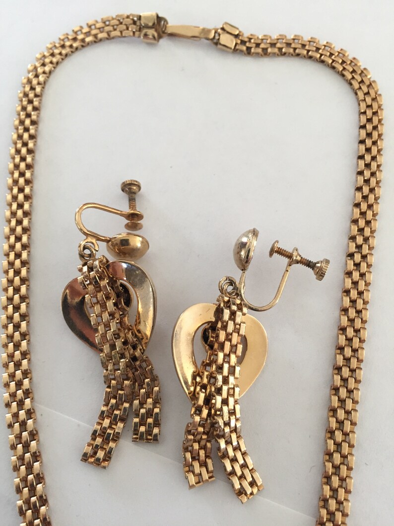 May include: A gold tone necklace and matching earrings with a heart-shaped pendant and dangling chains. The necklace is a box chain and the earrings are clip-on.