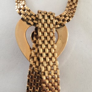 May include: A gold chain necklace with a heart-shaped pendant. The chain is made of small, square links. The pendant is a heart with a cutout in the center. The chain is tied in a bow at the bottom of the pendant.